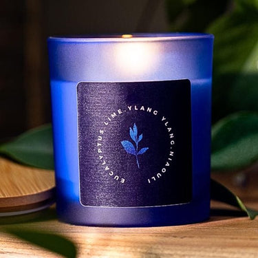 Blue candle with a label on a wooden surface with green leaves in the background